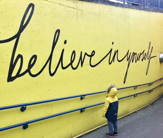 Toddler standing in front of wall with sign saying, “Believe in yourself”
